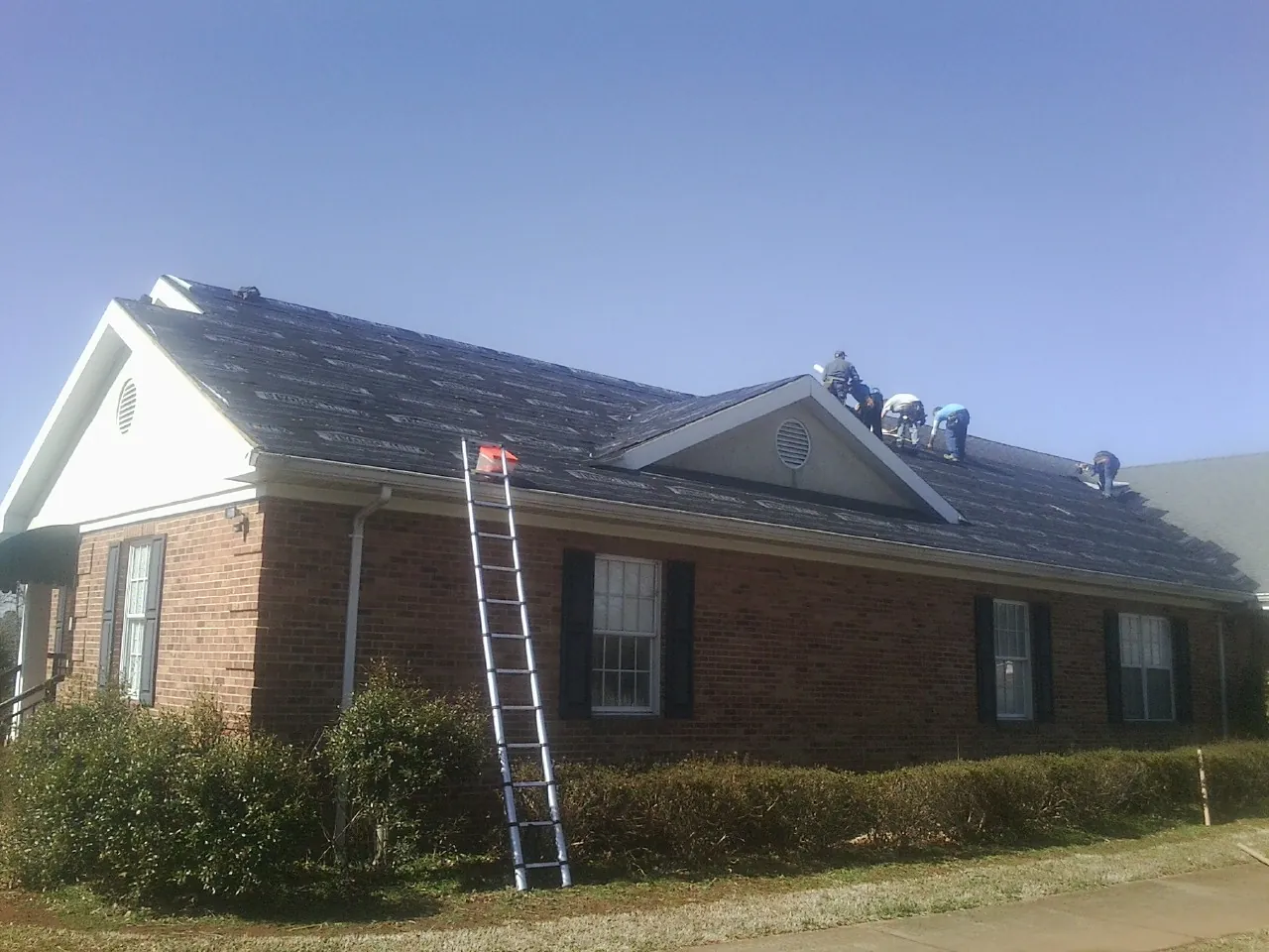 Crew tearing shingles off a brick ranch roof with a ladder set — Emergency Roof Repair work by our crew in Raymond